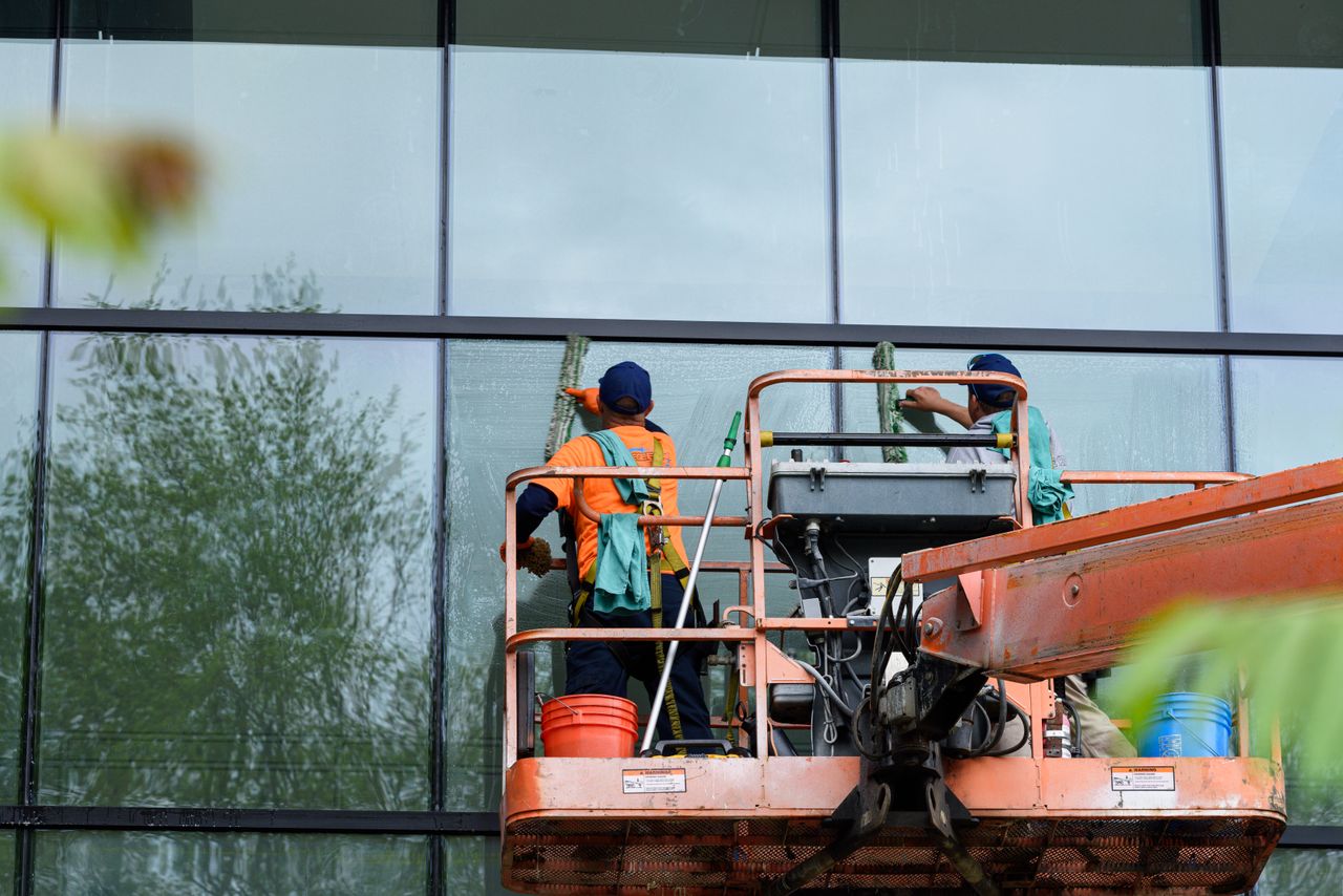 Deux ouvriers lavent les fenêtres d’une façade vitrée à l’aide d’une plateforme élévatrice, améliorant l’esthétique du bâtiment.